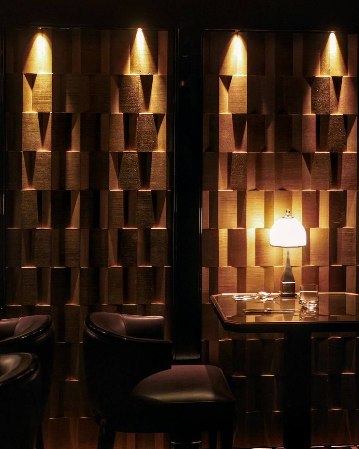 A lit lamp over a table and leather chairs against a dark timber wall